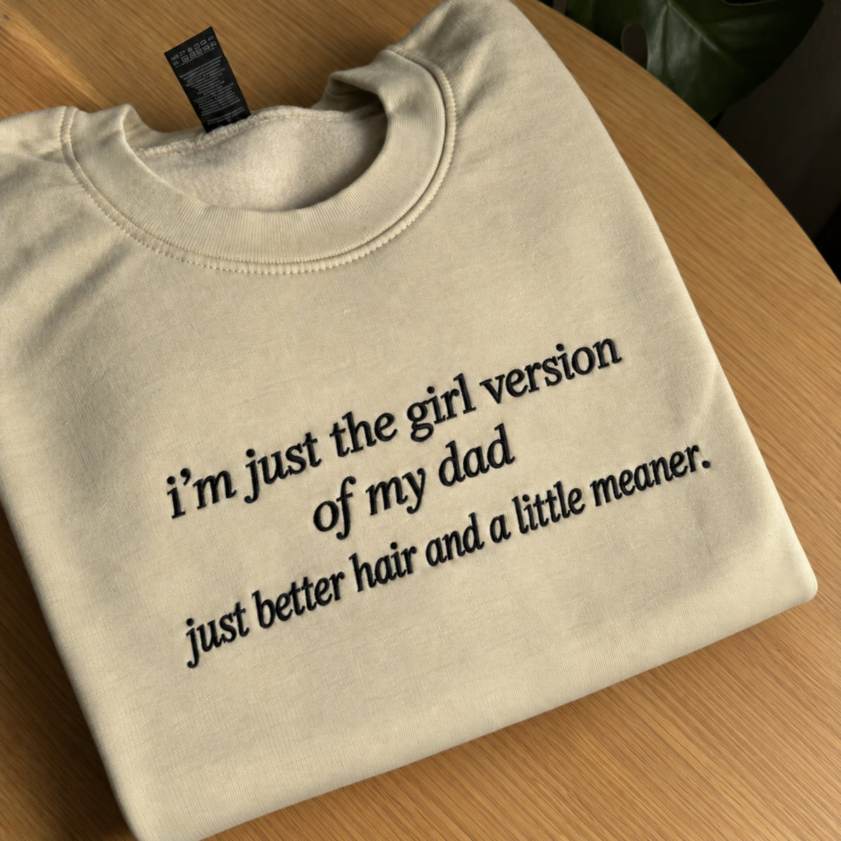 Cream embroidered sweatshirt folded on a wooden table, featuring black embroidered text that reads “I’m just the girl version of my dad, just better hair and a little meaner.”