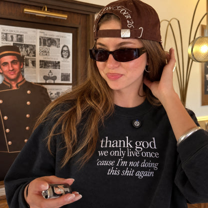 Woman wearing a black embroidered sweatshirt with the white quote “thank god we only live once ’cause I’m not doing this shit again,” styled with a brown cap and sunglasses in a casual indoor setting.