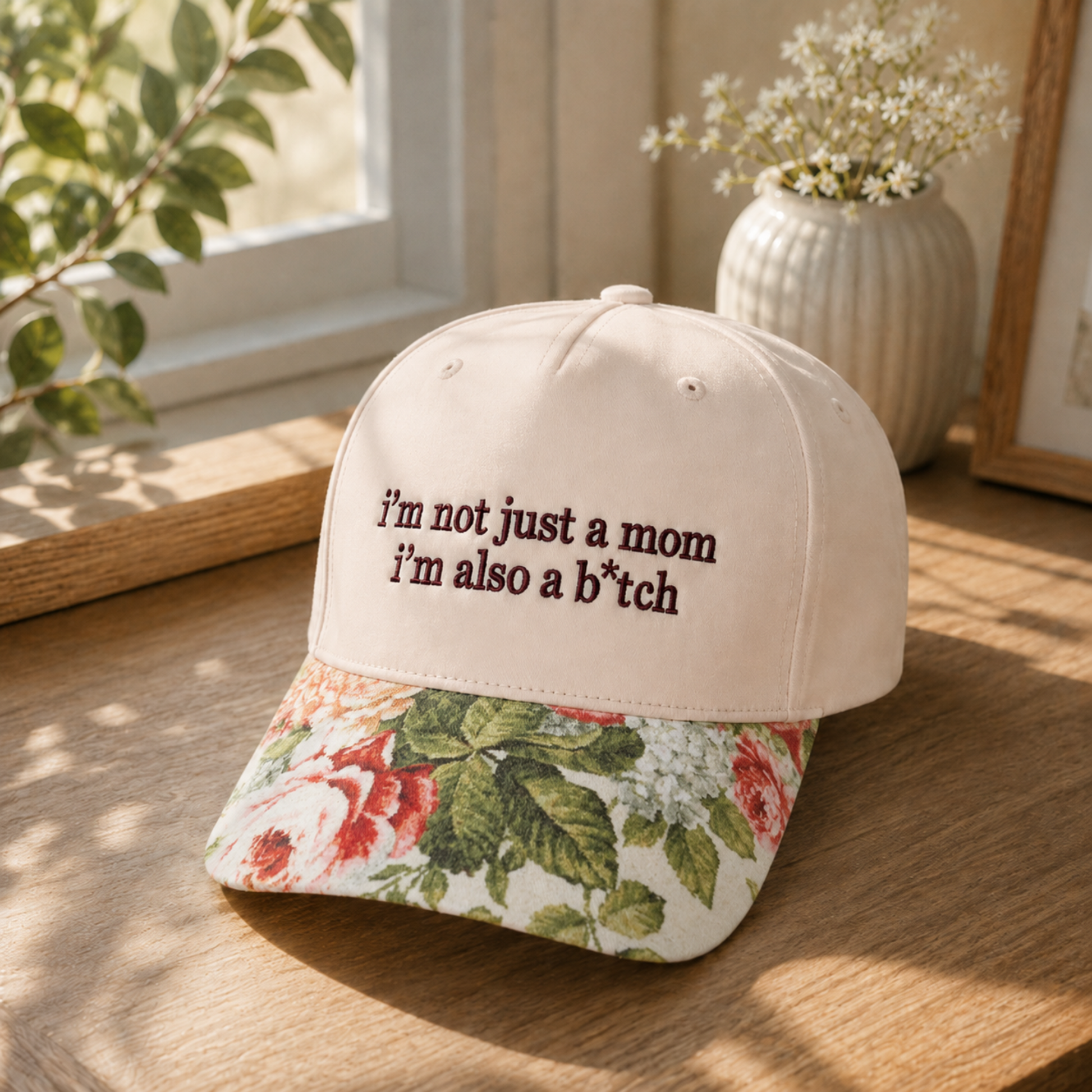 A baseball cap resting on a wooden table in a sunlit room. The cap has a light beige crown with dark brown embroidered text reading “i’m not just a mom i’m also a b*tch” and a floral brim with red, green, and white flowers.
