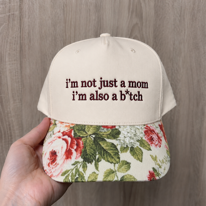 A hand holding a cream-colored baseball cap with dark brown embroidered text reading 'i'm not just a mom i'm also a b*tch' on the front, featuring a floral-patterned brim with red and pink roses and green leaves, set against a light wood background.