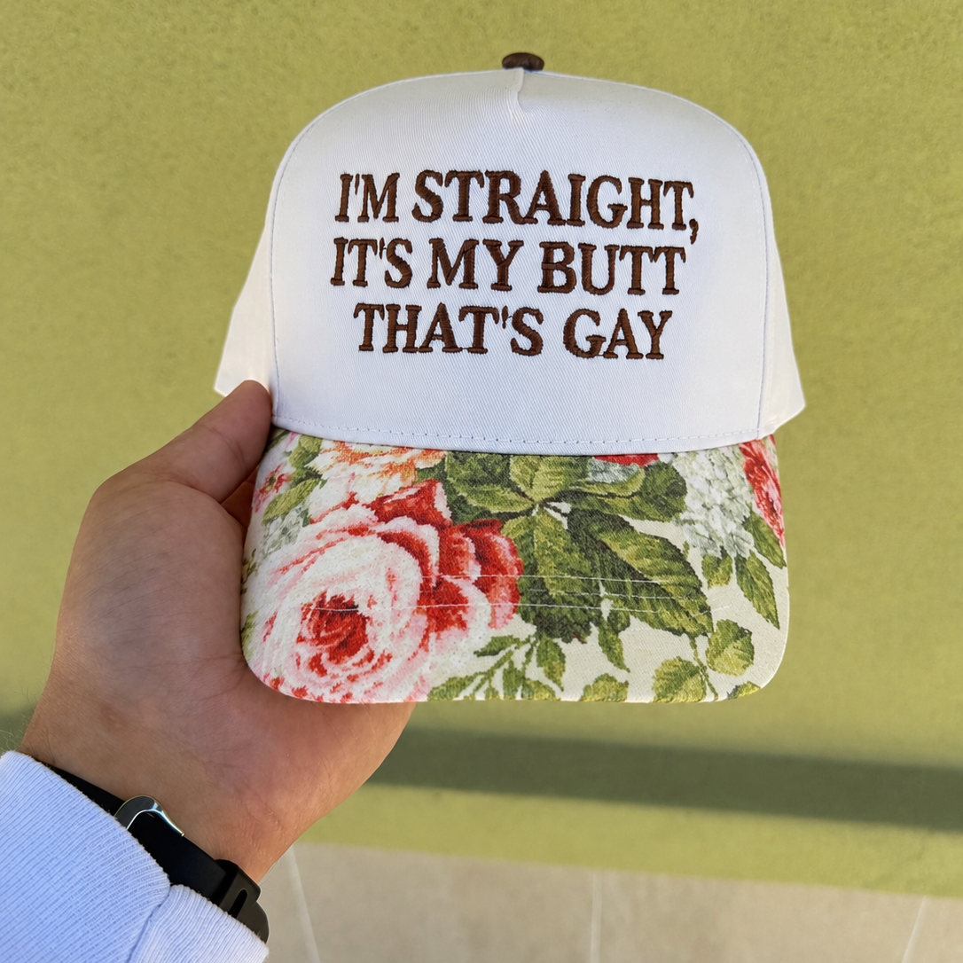 A hand holding a white trucker-style cap with brown text reading 'I'M STRAIGHT, IT'S MY BUTT THAT'S GAY' and a floral brim with red, pink, and green rose print, against a yellow-green wall.
