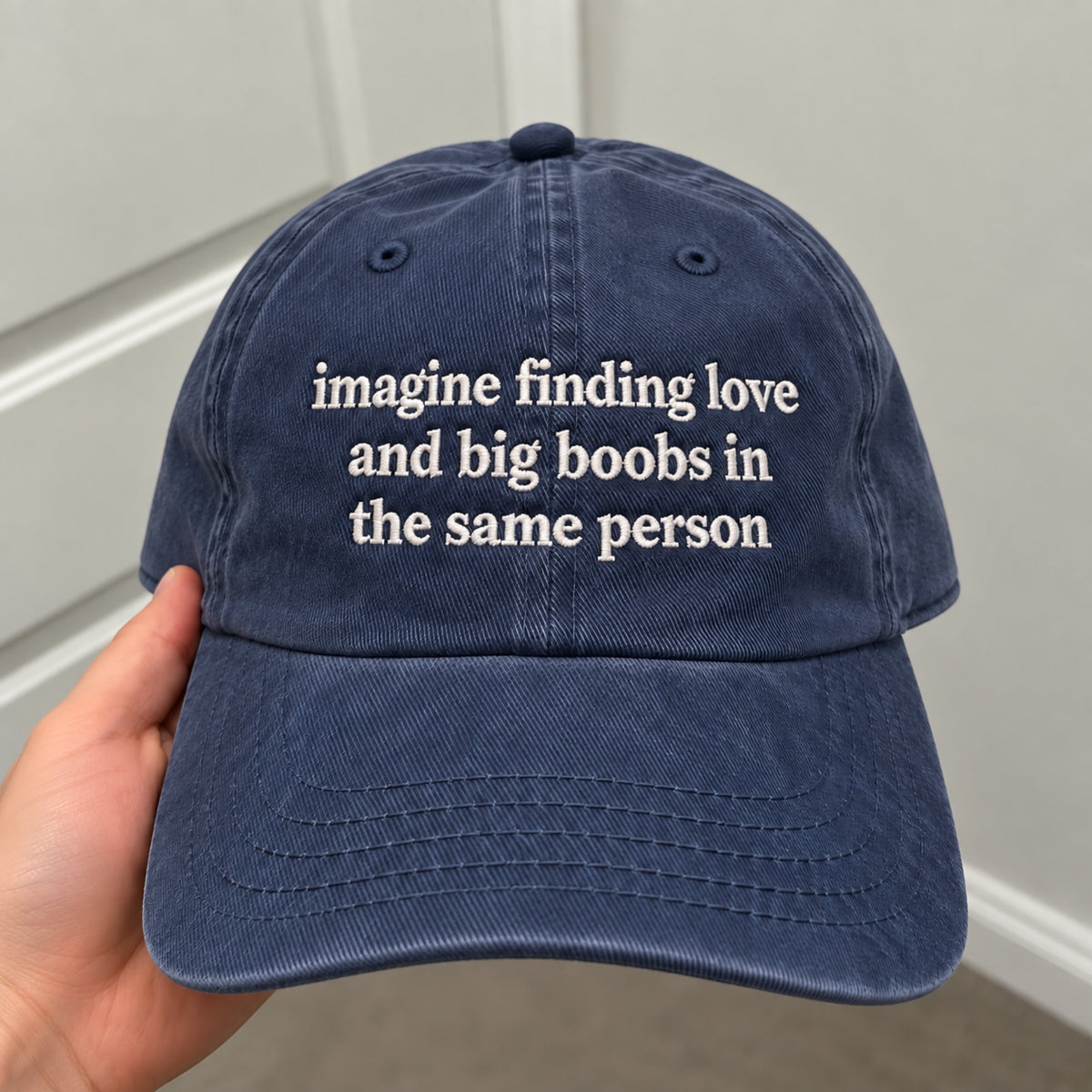 A navy blue washed cotton baseball cap held by a hand, featuring white embroidered text that reads 'imagine finding love and big boobs in the same person'.