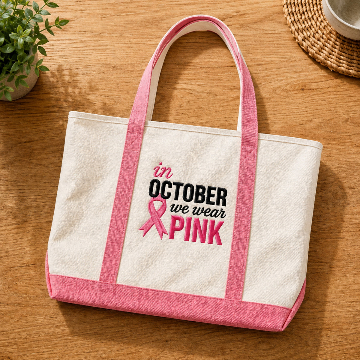 A cream canvas tote bag with pink handles, bottom trim, and side straps, featuring the embroidered text 'in OCTOBER we wear PINK' with a pink ribbon on the front, displayed on a wooden tabletop.