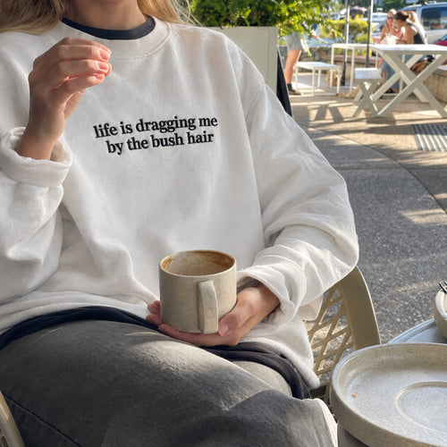A person sitting outdoors at a cafe wearing a white crewneck sweatshirt with the black printed text 'life is dragging me by the bush hair'. The person is holding a beige ceramic mug.