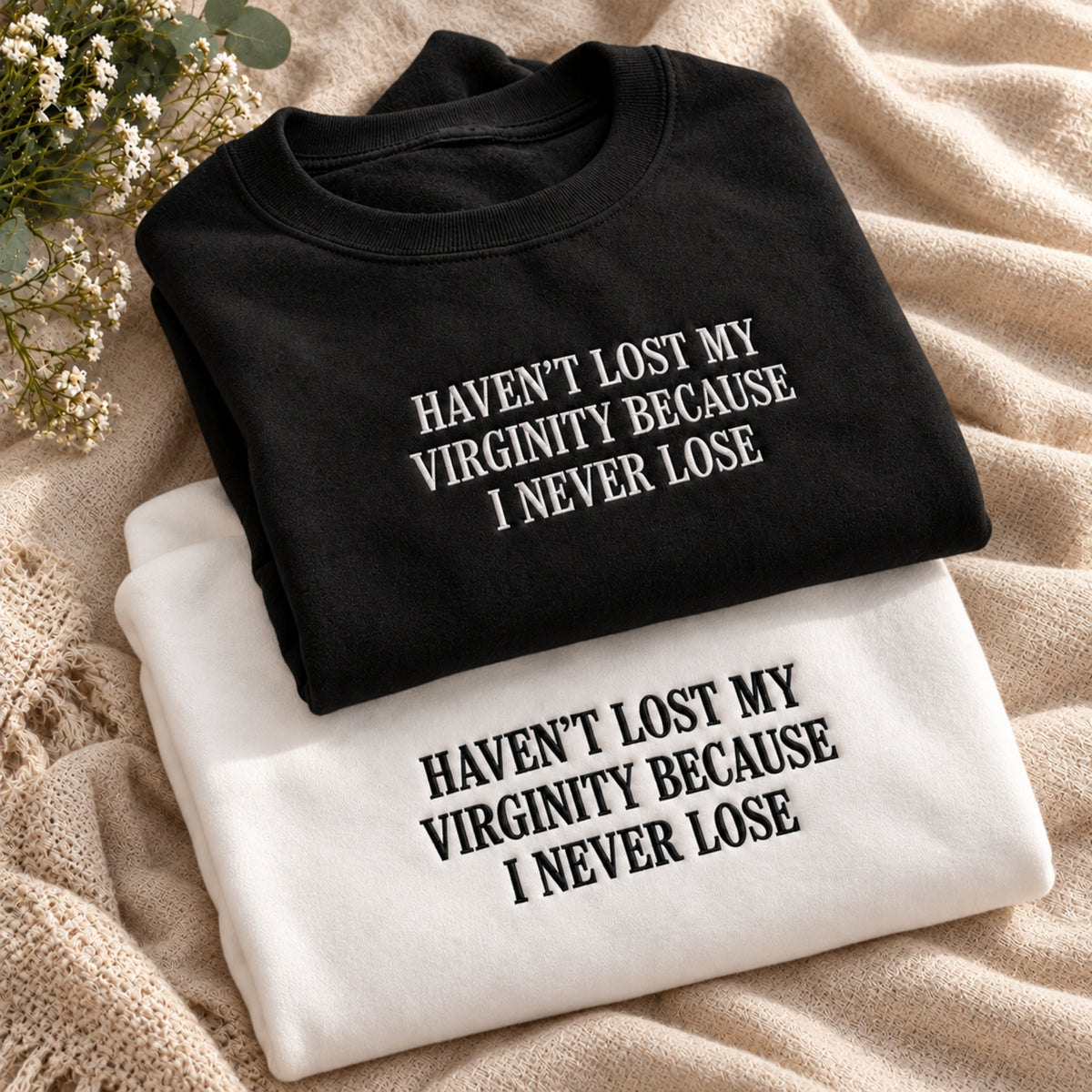 Two folded graphic T-shirts displayed on a beige textured blanket with small white flowers. The top shirt is black with white text reading 'HAVEN'T LOST MY VIRGINITY BECAUSE I NEVER LOSE', and the bottom shirt is white with black text reading the same phrase.