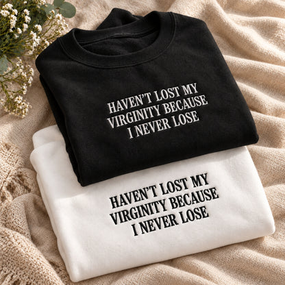 Two folded graphic T-shirts displayed on a beige textured blanket with small white flowers. The top shirt is black with white text reading 'HAVEN'T LOST MY VIRGINITY BECAUSE I NEVER LOSE', and the bottom shirt is white with black text reading the same phrase.