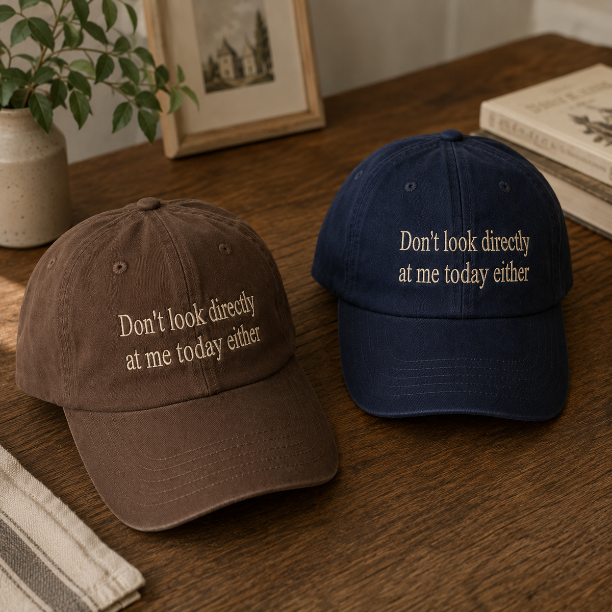 Two embroidered baseball caps on a wooden table: one brown and one navy blue, both with the phrase 'Don't look directly at me today either' in light beige lettering. A plant, framed picture, and books are visible in the background.
