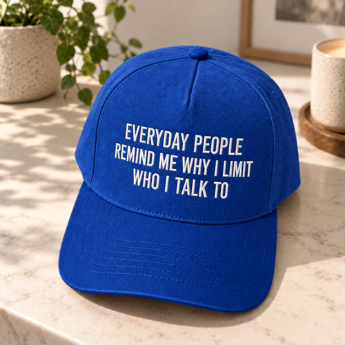 A royal blue baseball cap with white embroidered text reading 'EVERYDAY PEOPLE REMIND ME WHY I LIMIT WHO I TALK TO' sits on a light marble surface with a potted plant and ceramic cup in the background.