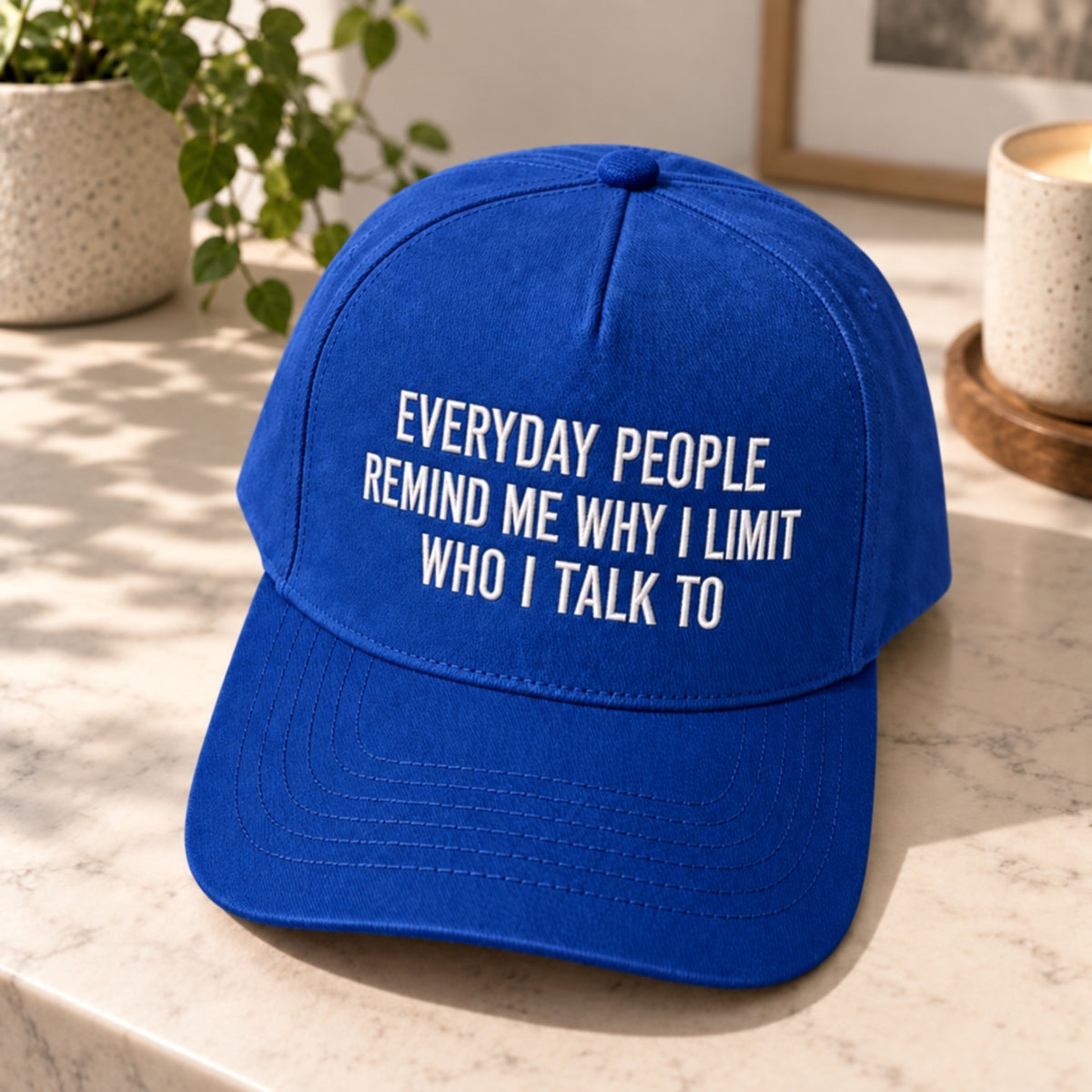 A royal blue baseball cap with white embroidered text reading 'EVERYDAY PEOPLE REMIND ME WHY I LIMIT WHO I TALK TO' sits on a light marble surface with a potted plant and ceramic cup in the background.