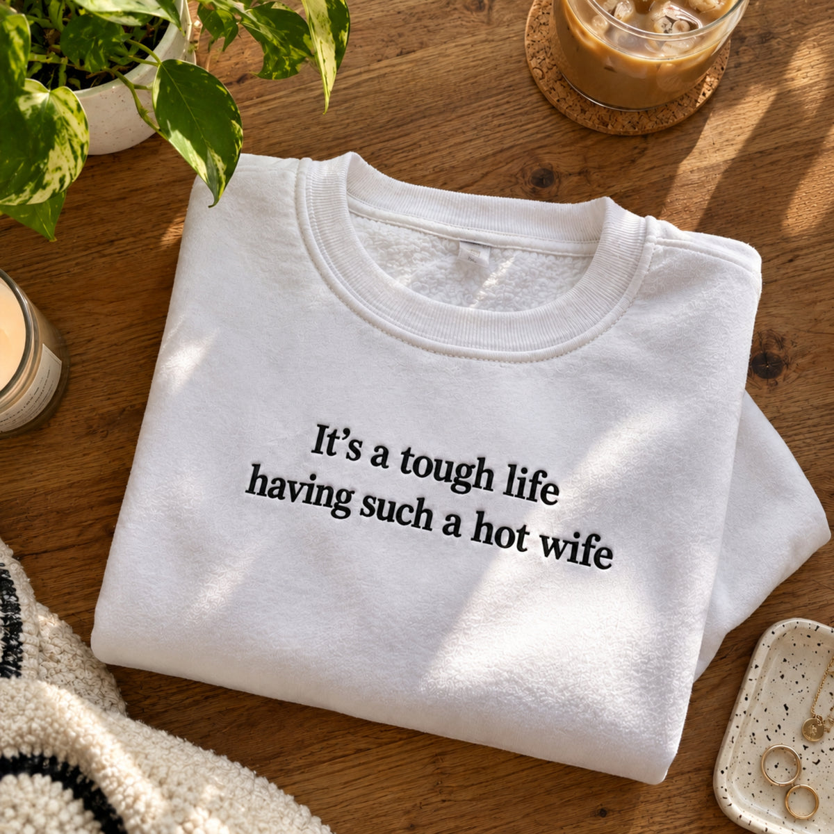 A folded white crewneck sweatshirt with black text reading 'It's a tough life having such a hot wife' on a wooden table, surrounded by lifestyle props like a plant, an iced coffee, and a candle.