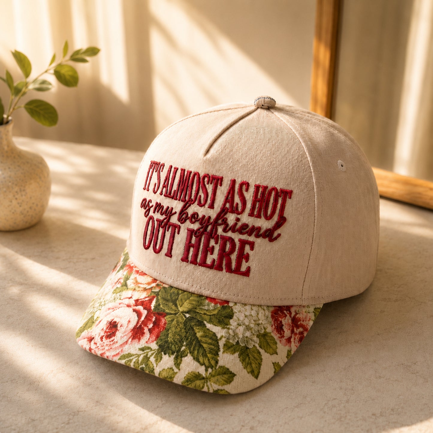 A beige baseball cap with red embroidered text reading 'IT'S ALMOST AS HOT as my boyfriend OUT HERE' and a floral-patterned visor, styled on a tabletop with warm sunlight and a small plant in the background.