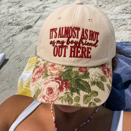 A person at the beach wearing a beige baseball cap with red embroidered text reading 'IT'S ALMOST AS HOT as my boyfriend OUT HERE' and a floral visor with pink roses and green leaves.