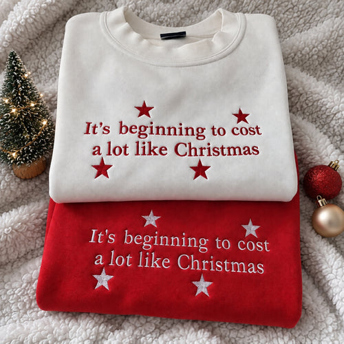 Two folded Christmas-themed sweatshirts stacked on a soft white fleece blanket, with a small lit tree and holiday ornaments as props. The top sweatshirt is off-white with red embroidered text reading 'It's beginning to cost a lot like Christmas' and red stars.