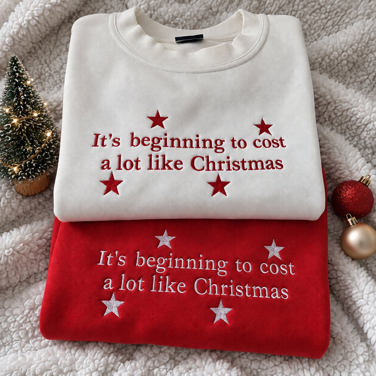 Two folded Christmas-themed sweatshirts stacked on a soft white fleece blanket, with a small lit tree and holiday ornaments as props. The top sweatshirt is off-white with red embroidered text reading 'It's beginning to cost a lot like Christmas' and red stars.