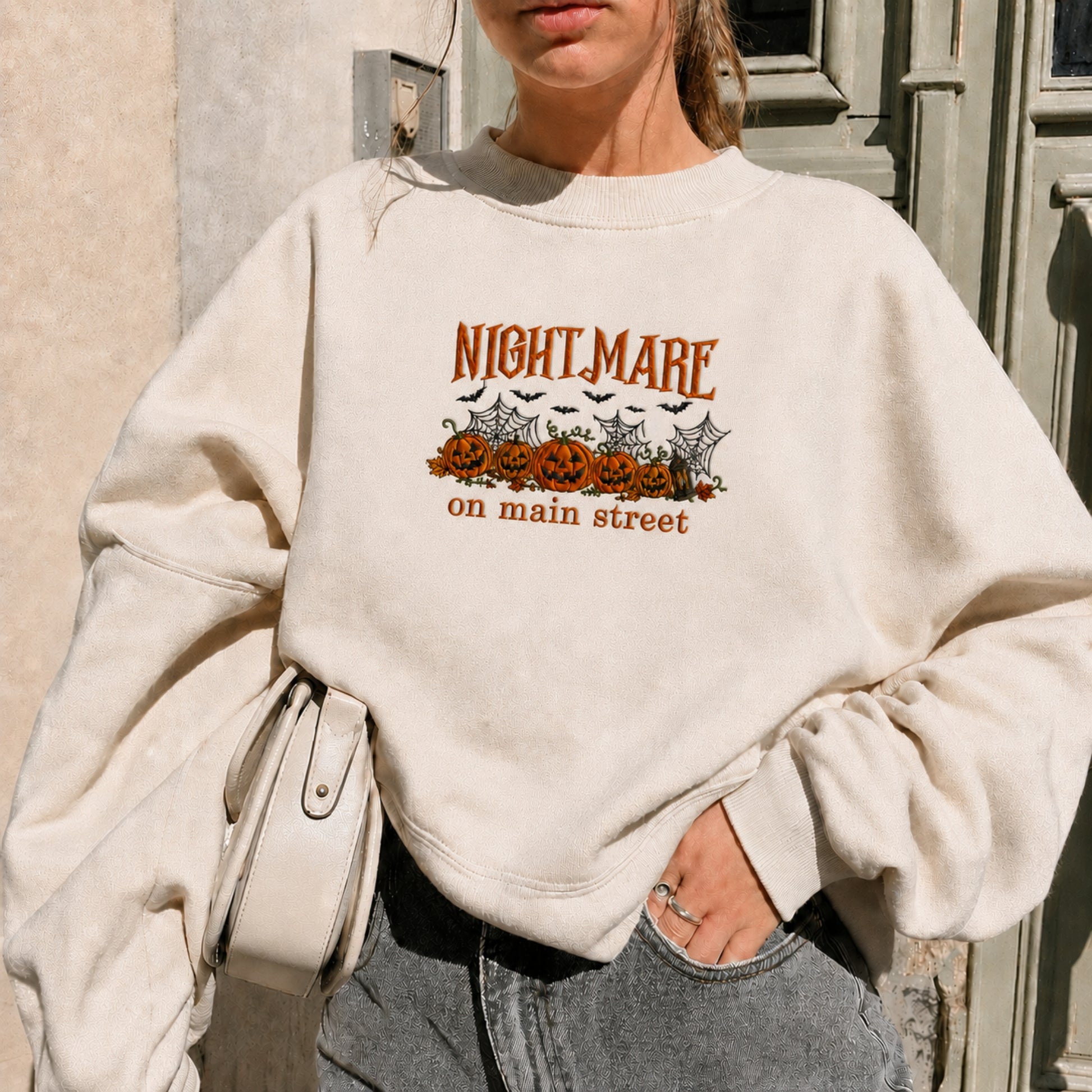 A woman wearing an oversized cream-colored sweatshirt with a Halloween graphic that reads 'NIGHTMARE on main street' above jack-o'-lanterns, bats, and cobwebs. She is also wearing gray jeans and holding a light-colored shoulder bag.