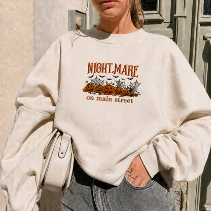 A woman wearing an oversized cream-colored sweatshirt with a Halloween graphic that reads 'NIGHTMARE on main street' above jack-o'-lanterns, bats, and cobwebs. She is also wearing gray jeans and holding a light-colored shoulder bag.