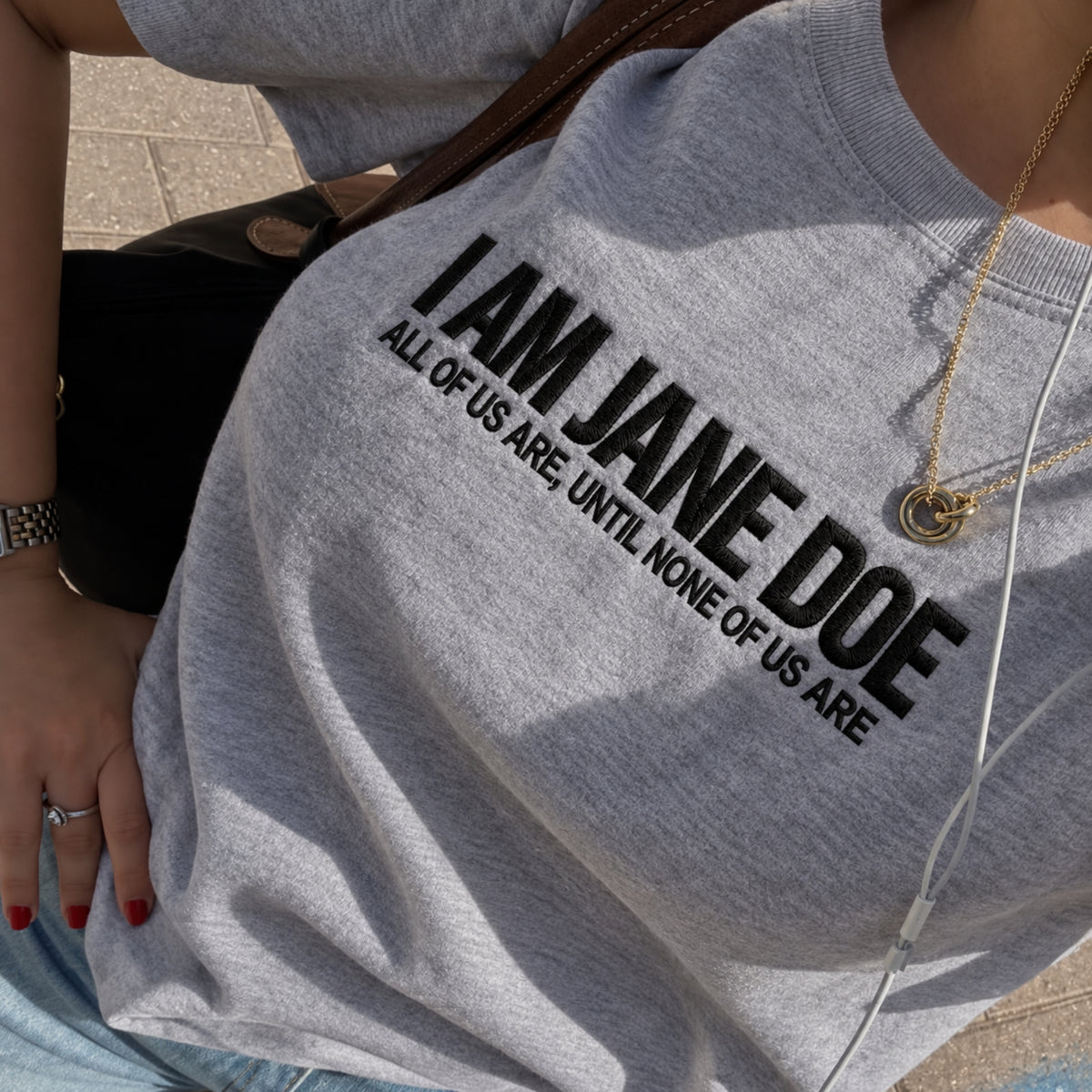 A close-up of a person wearing a heather gray crewneck sweatshirt with bold black text reading 'I AM JANE DOE' and smaller text 'ALL OF US ARE, UNTIL NONE OF US ARE'.