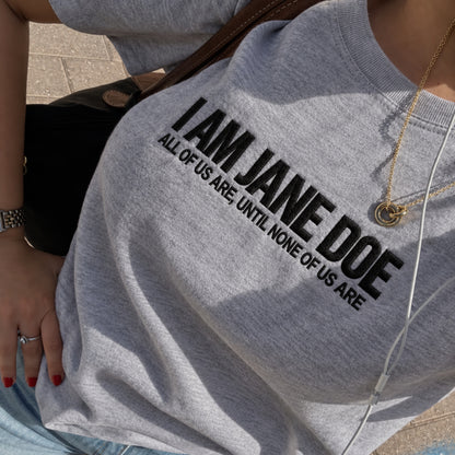A close-up of a person wearing a heather gray crewneck sweatshirt with bold black text reading 'I AM JANE DOE' and smaller text 'ALL OF US ARE, UNTIL NONE OF US ARE'.