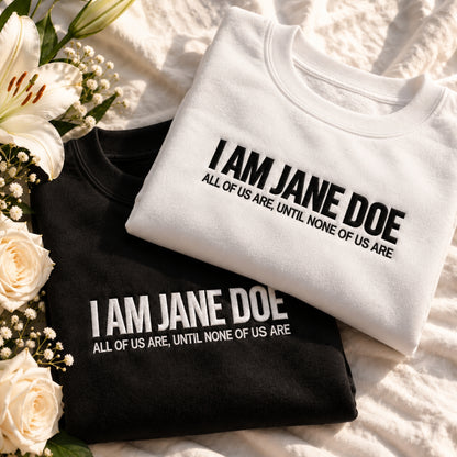 Two folded graphic T-shirts, one white and one black, displayed on a textured cream fabric with white roses and lilies. The shirts feature the text 'I AM JANE DOE' and 'ALL OF US ARE, UNTIL NONE OF US ARE' in contrasting black and white lettering.
