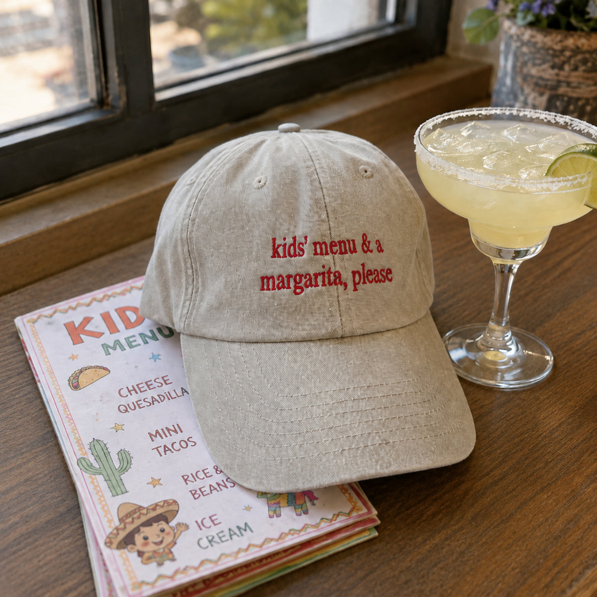 A beige baseball cap with red embroidered text reading 'kids' menu & a margarita, please' sits on a wooden table next to a margarita glass and a kids' menu.