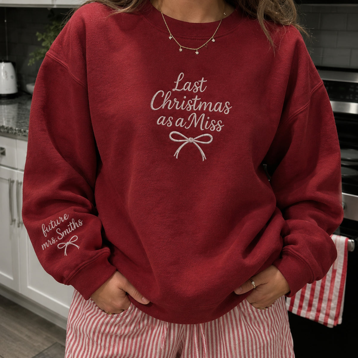 A person wearing a dark red crewneck sweatshirt with white script text reading 'Last Christmas as a Miss' and 'future miss Smiths' on the sleeve, paired with pink and white striped pajama pants in a kitchen setting.