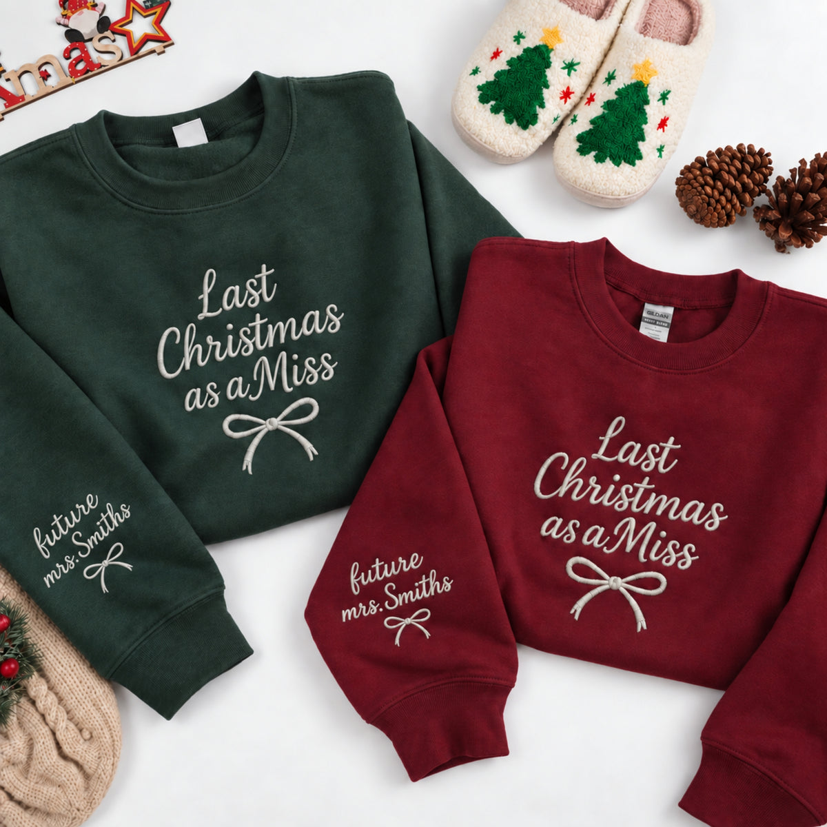 Two Christmas-themed crewneck sweatshirts laid flat on a white background, one dark green and one burgundy, both with white embroidered text reading 'Last Christmas as a Miss' and a bow graphic. Additional holiday props including slippers, pinecones, and a knit hat are arranged around them.