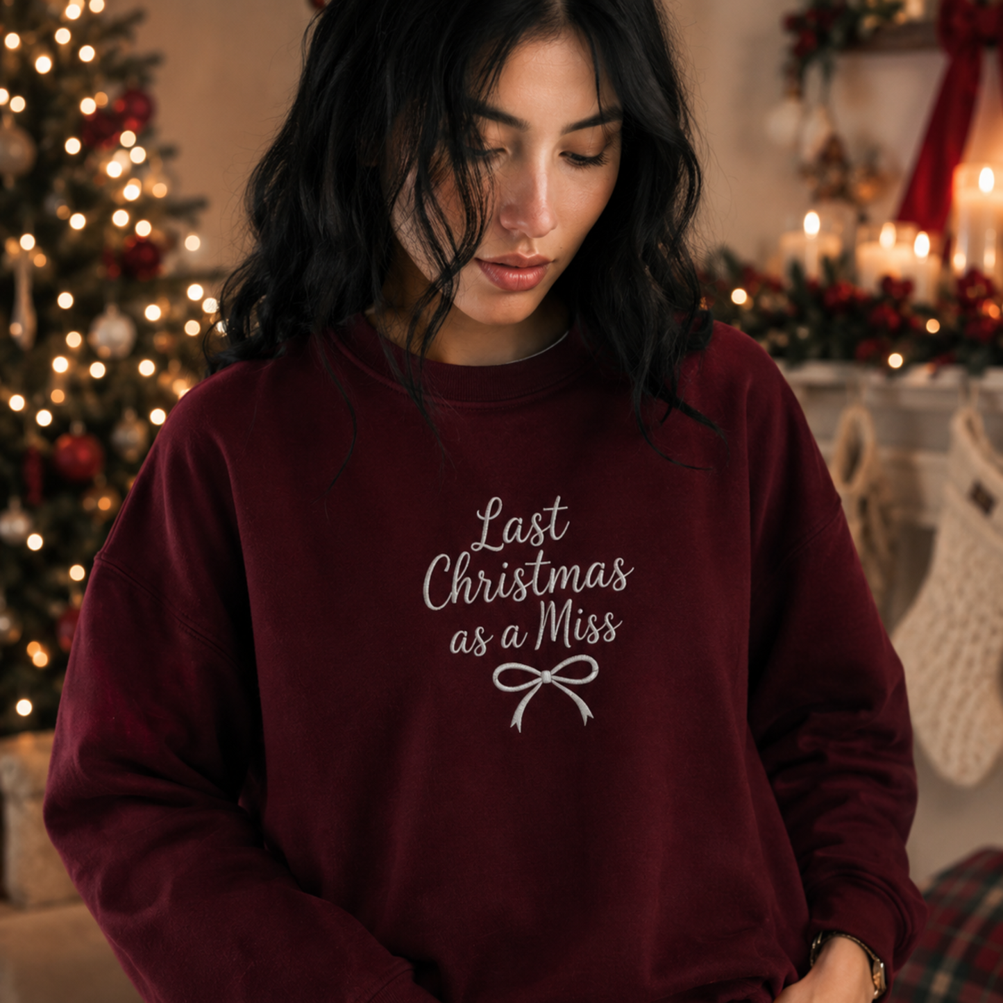 Woman wearing a maroon crewneck sweatshirt embroidered with white text reading "Last Christmas as a Miss" and a ribbon bow, set in a cozy holiday Christmas decor scene.