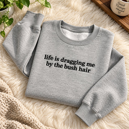 A light heather gray crewneck sweatshirt with the phrase 'life is dragging me by the bush hair' printed in black across the chest, styled on a cream faux fur rug with a knit blanket and green plant leaves.