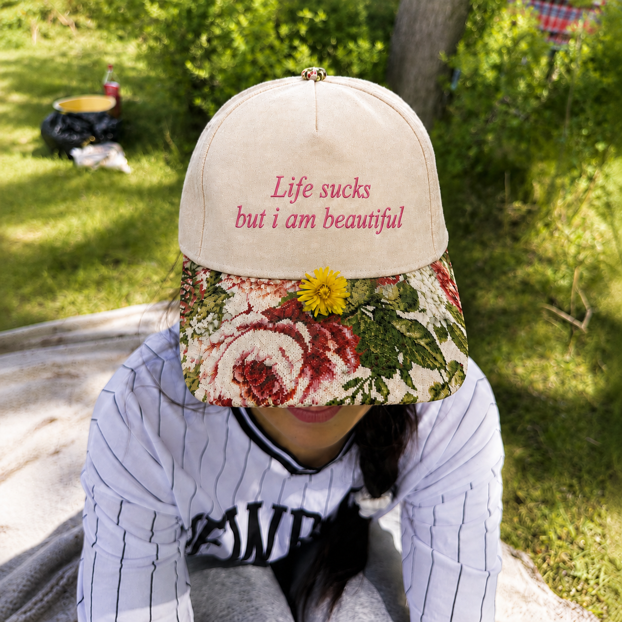 A person outdoors wearing a beige baseball cap with pink embroidered text reading 'Life sucks but i am beautiful' and a floral brim with a yellow flower accent, paired with a white pinstripe shirt.
