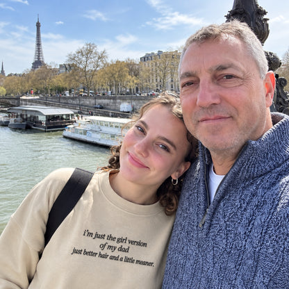 A father and daughter posing together by the Seine River in Paris, with the Eiffel Tower in the background. The daughter is wearing a beige sweatshirt featuring black embroidered text that reads, “I’m just the girl version of my dad, just better hair and a little meaner.”