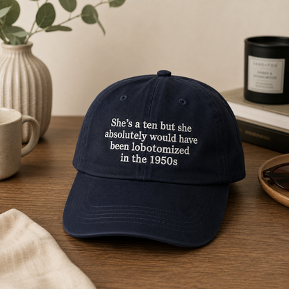 A navy blue baseball cap with white embroidered text reading 'She's a ten but she absolutely would have been lobotomized in the 1950s' displayed on a wooden table with lifestyle props.
