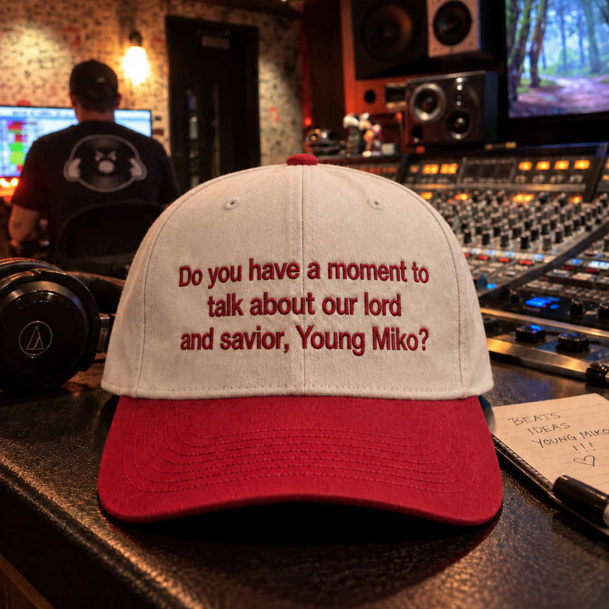 A baseball cap with a white crown and red brim, featuring the text 'Do you have a moment to talk about our lord and savior, Young Miko?' in red embroidery, displayed on a mixing board in a recording studio.