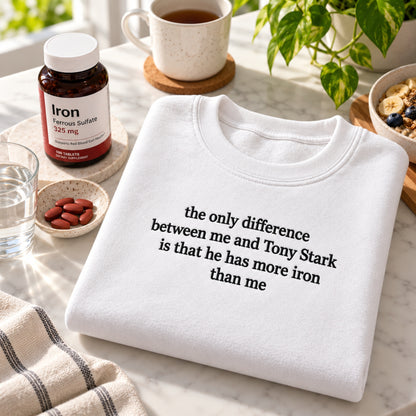 A folded white crewneck sweatshirt with black text reading 'the only difference between me and Tony Stark is that he has more iron than me' sits on a marble surface. The scene includes an Iron supplement bottle, a mug, a glass of water, and a plant in a bright, styled setting.