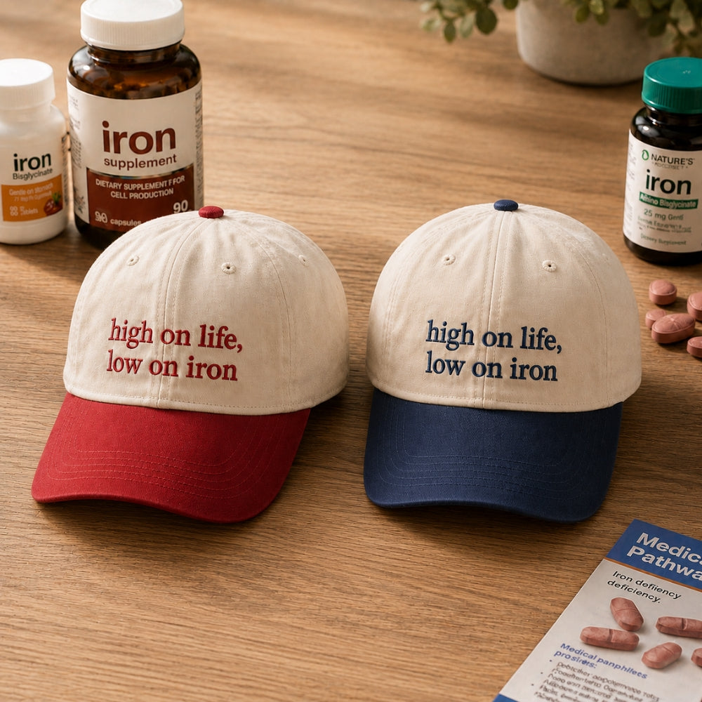 Two baseball caps displayed on a wooden tabletop, surrounded by supplement bottles and pills. The left cap has a red brim and cream crown with red embroidered text reading 'high on life, low on iron'.