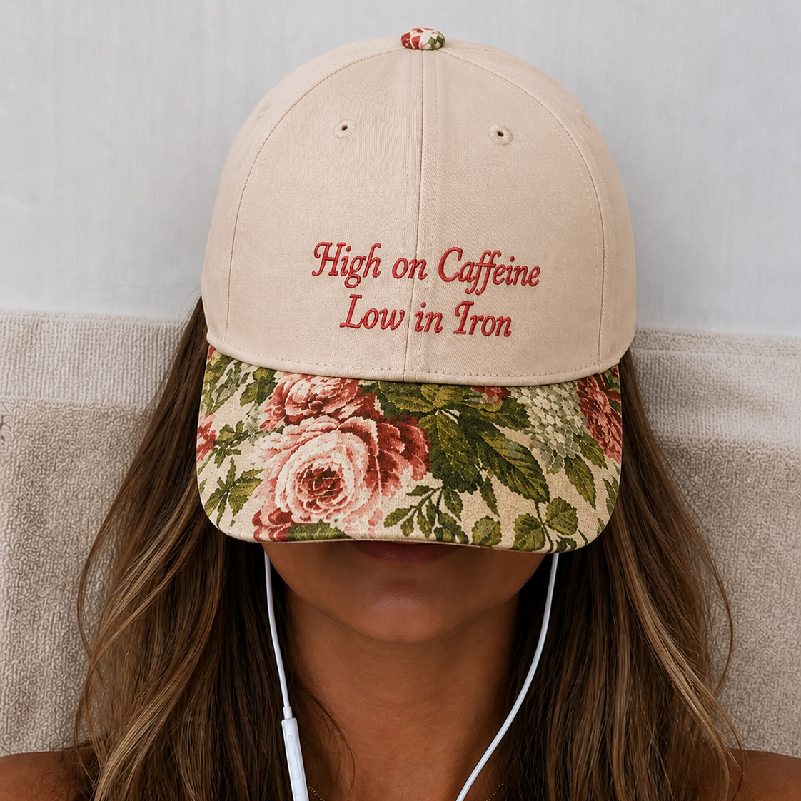 Woman with brown hair wearing a beige baseball cap with red embroidered text 'High on Caffeine Low in Iron', a vintage floral rose tapestry visor, and wired earphones.