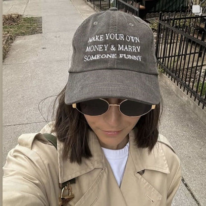 A person taking a selfie outdoors wearing a dark gray corduroy baseball cap with white embroidered text that reads 'MAKE YOUR OWN MONEY & MARRY SOMEONE FUNNY', along with sunglasses, a beige trench coat, and a white shirt.