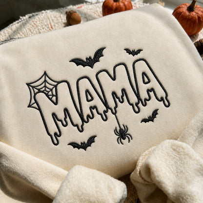 A cream-colored fleece blanket with black embroidered 'MAMA' lettering in a dripping font, surrounded by Halloween motifs like bats, a spider, and a spiderweb. Small pumpkins and acorns are visible in the background.