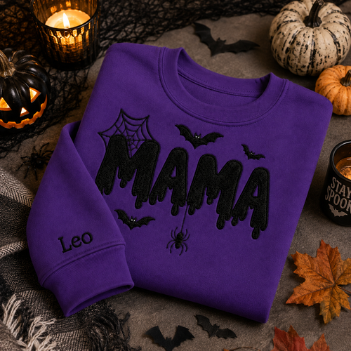 Vibrant purple Halloween crewneck sweatshirt featuring black embroidered "MAMA" lettering with cobwebs, bats, a dangling spider, and custom name "Leo" embroidered on the sleeve, styled on a flat lay with jack-o'-lanterns, pumpkins, and a candle.