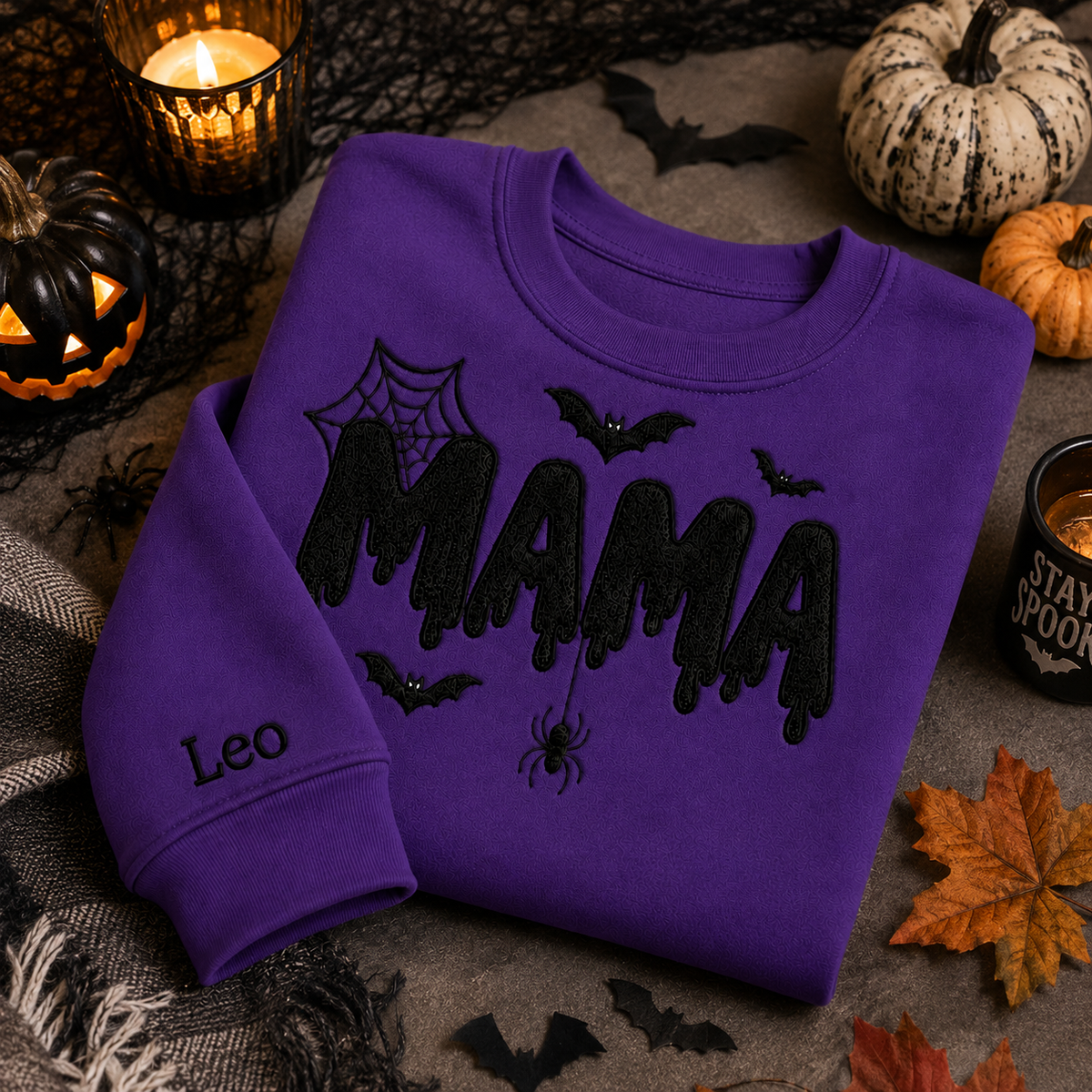 Vibrant purple Halloween crewneck sweatshirt featuring black embroidered "MAMA" lettering with cobwebs, bats, a dangling spider, and custom name "Leo" embroidered on the sleeve, styled on a flat lay with jack-o'-lanterns, pumpkins, and a candle.