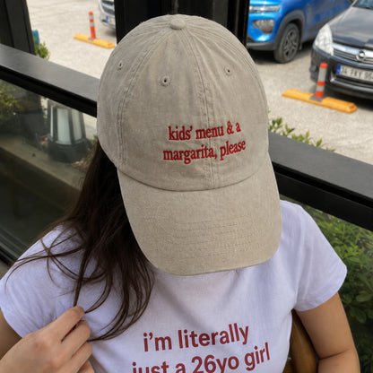 A person wearing a beige baseball cap with red embroidered text reading 'kids' menu & a margarita, please' and a white graphic T-shirt in an outdoor setting.