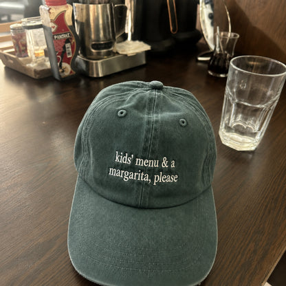 A dark green baseball cap with the phrase 'kids' menu & a margarita, please' embroidered in white on the front, resting on a dark wooden table with a glass and coffee equipment in the background.