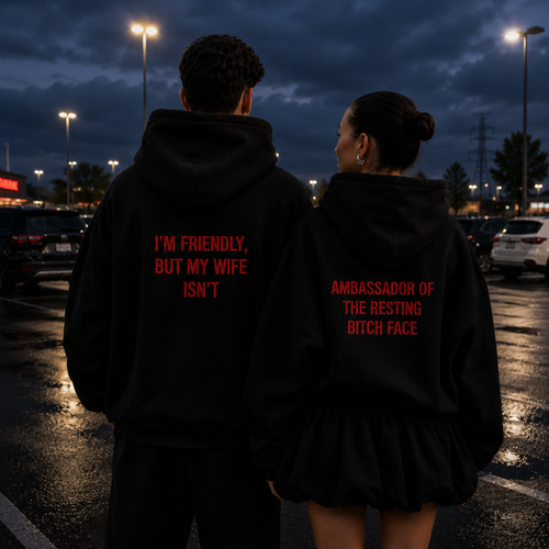 Two people standing in a parking lot at dusk, seen from behind, wearing matching black hoodies with red text. The hoodie on the left reads 'I'M FRIENDLY, BUT MY WIFE ISN'T' and the hoodie on the right reads 'AMBASSADOR OF THE RESTING BITCH FACE'.
