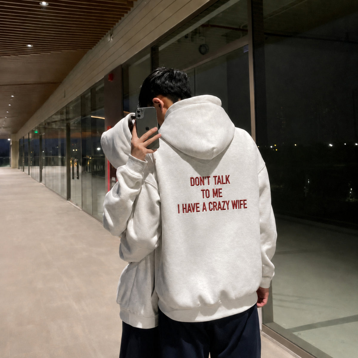 A person standing in a modern walkway takes a mirror selfie while wearing a light gray hoodie from the back, which features the red text 'DON'T TALK TO ME I HAVE A CRAZY WIFE'.