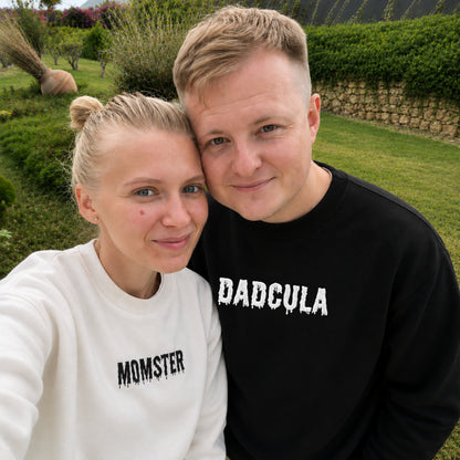 A man and a woman posing together outdoors in a garden setting, wearing matching sweatshirts with the words 'DADCULA' and 'MOMSTER' printed in white.