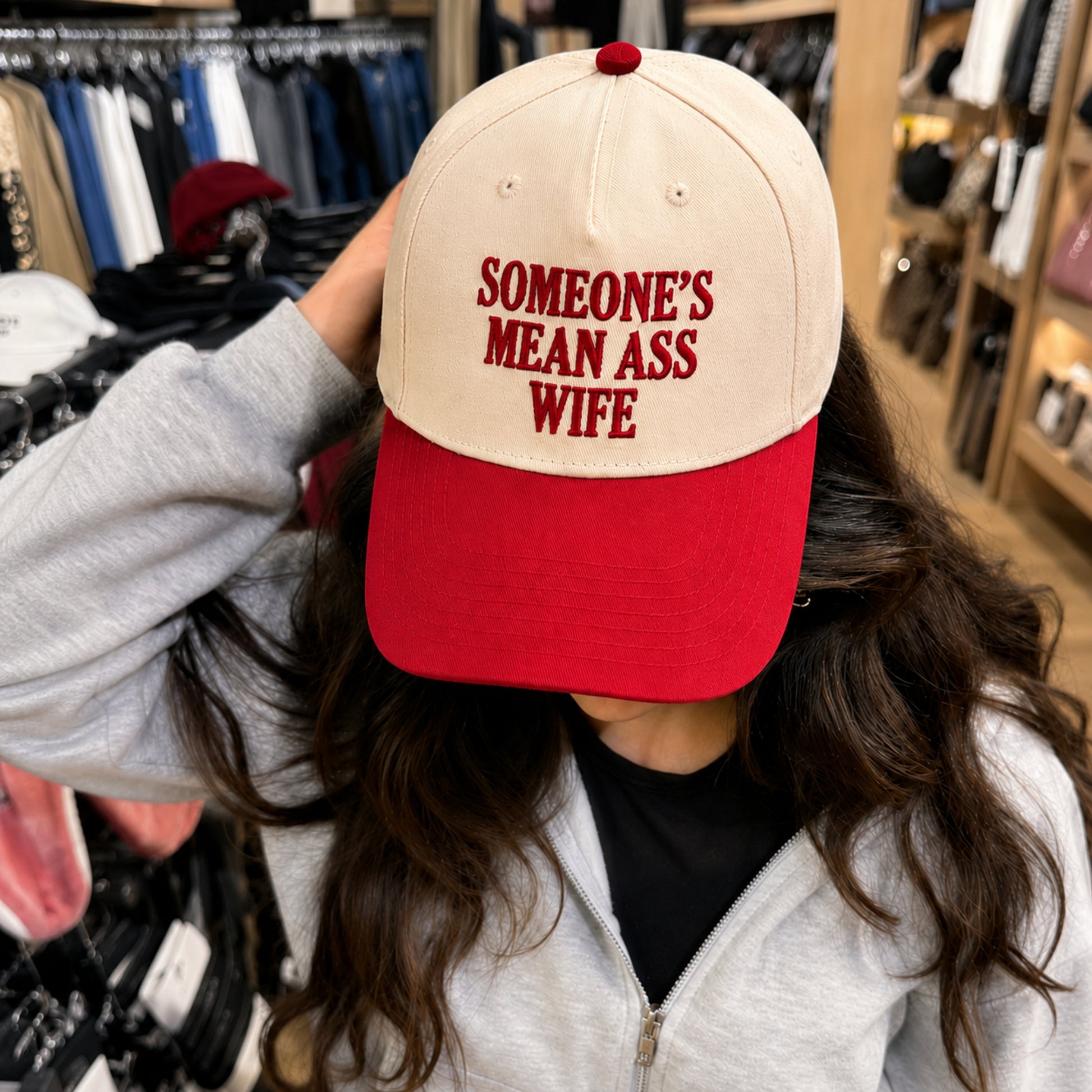 A person in a clothing store wearing a baseball cap with a cream-colored crown and a red visor. The cap features red embroidered text that reads 'SOMEONE'S MEAN ASS WIFE'.