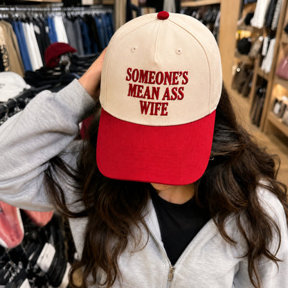 A person in a clothing store wearing a baseball cap with a cream-colored crown and a red visor. The cap features red embroidered text that reads 'SOMEONE'S MEAN ASS WIFE'.