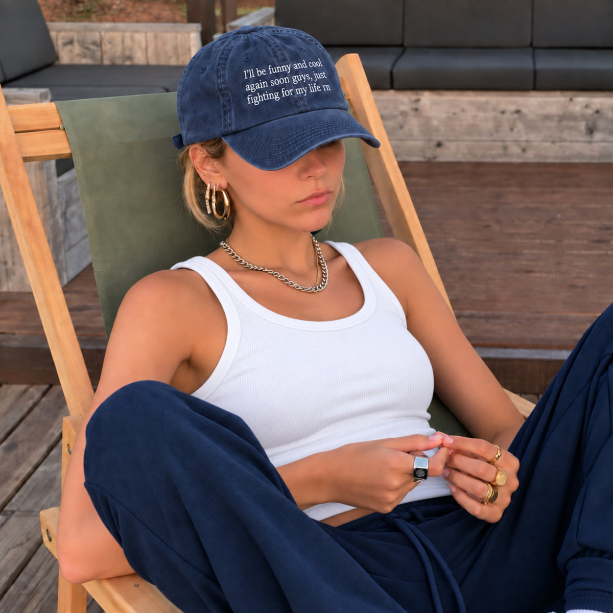 A woman sitting in a wooden deck chair wearing a navy blue baseball cap with white embroidered text, a white tank top, and navy pants.