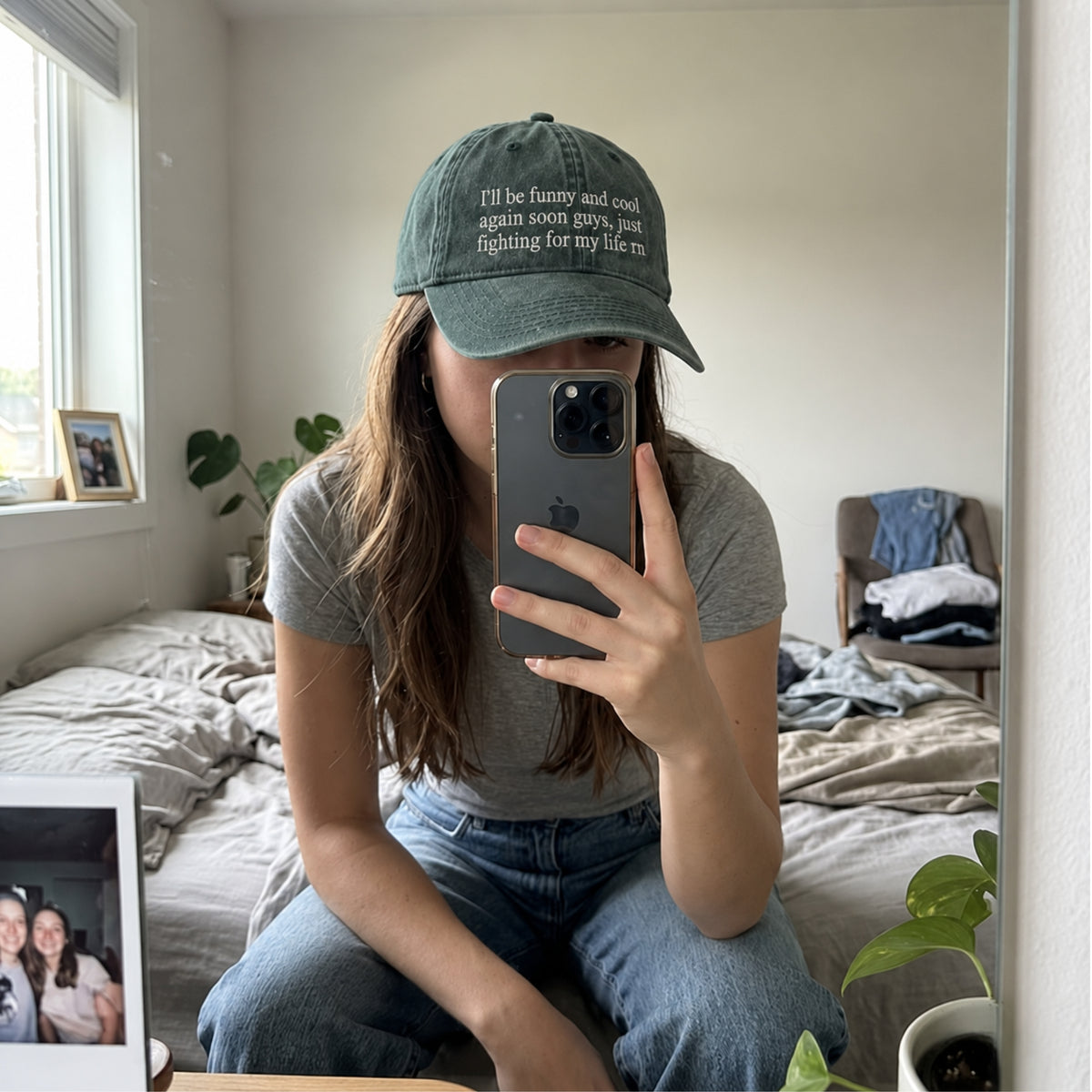 A person taking a mirror selfie in a bedroom, wearing a dark green baseball cap with the embroidered text 'I'll be funny and cool again soon guys, just fighting for my life.' and a gray t-shirt.