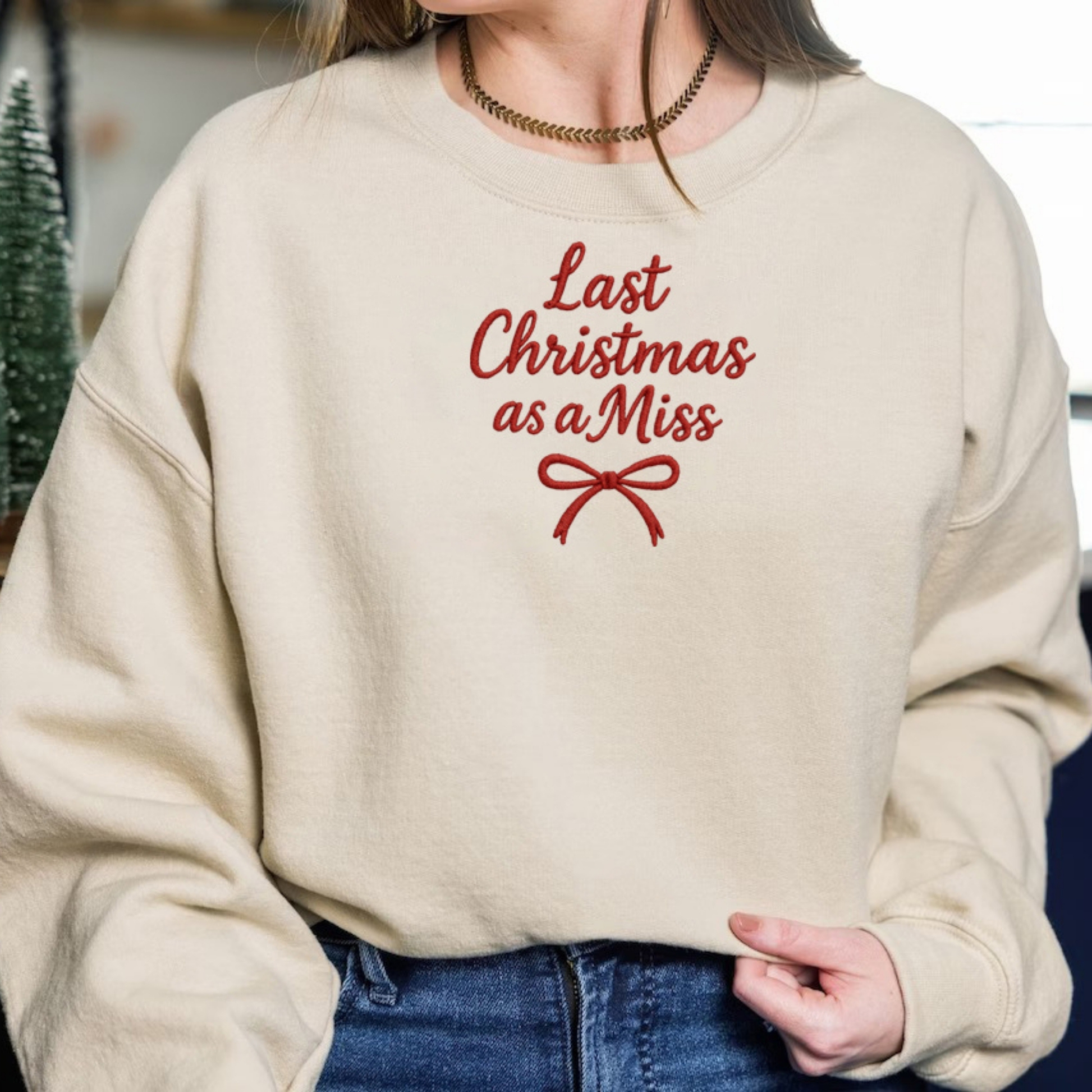 Beige crewneck sweatshirt embroidered with red text reading "Last Christmas as a Miss" and a red ribbon bow, set in a cozy holiday Christmas decor scene.