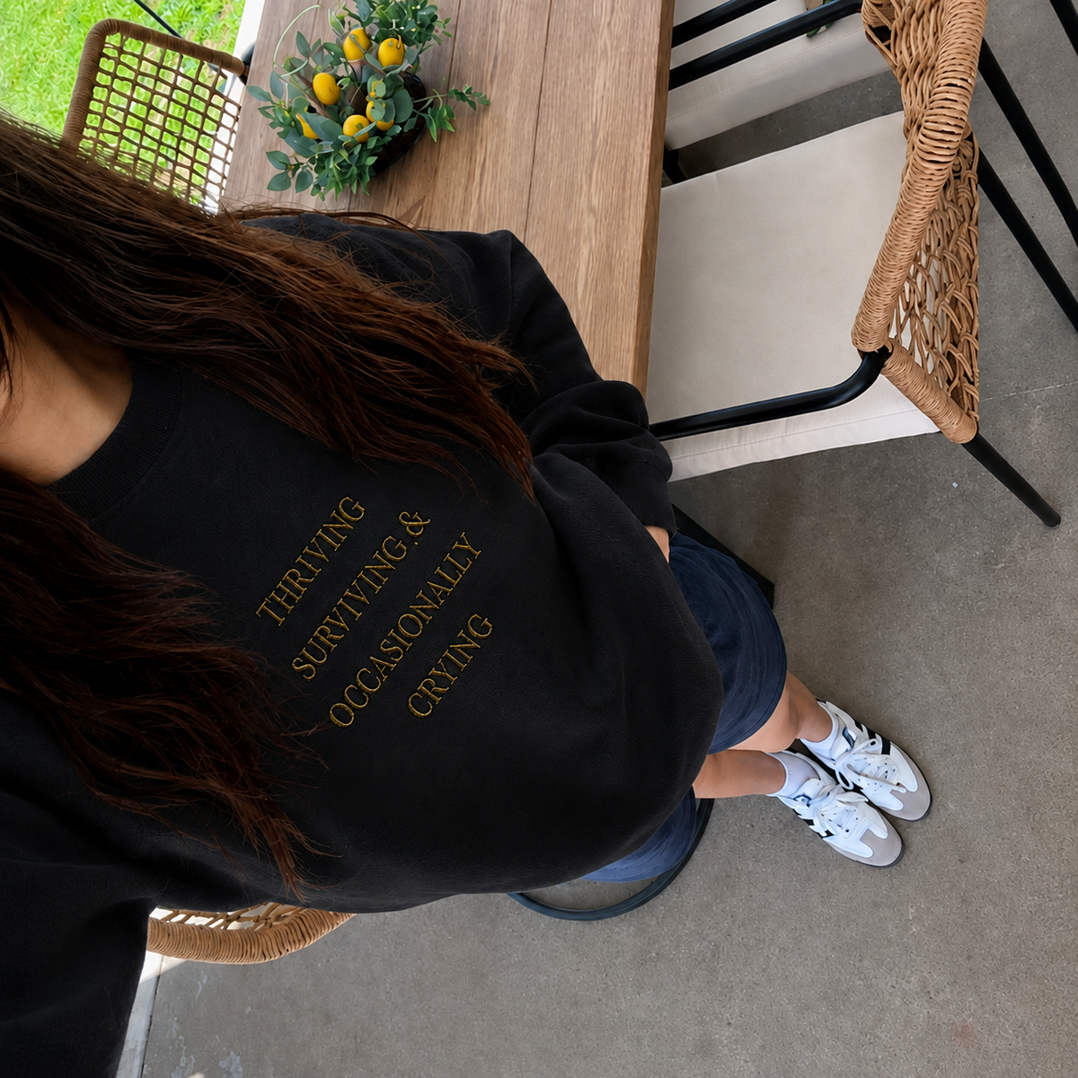 A person wearing a black crewneck sweatshirt with gold text that reads 'THRIVING SURVIVING & OCCASIONALLY CRYING' paired with blue shorts and white sneakers, photographed from an overhead angle in an outdoor patio setting.